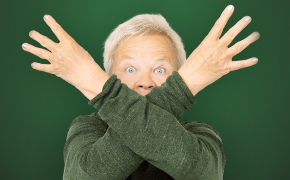 Vanha nainen seisoo vihreää taustaa vasten ja katsoo suoraan kameraan. Hän on ristinyt käsivartensa kasvojen eteen ja pitelee ylhäällä kahdeksaa sormea.